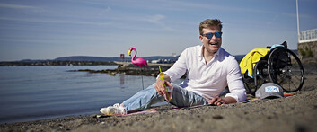 Jesper ligger på stranden og holder en drink. Han har solbriller på, og i bakgrunnen ser vi rullestolen og en rosa flamingo av plast.