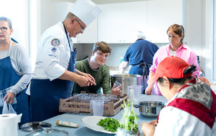 Kokk gir instrukser til tre kursdeltakere på et stort kjøkken.