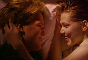 Ung mann og kvinne ligger i dobbeltseng og smiler til hverandre, kvinnen stryker nakken til mannen