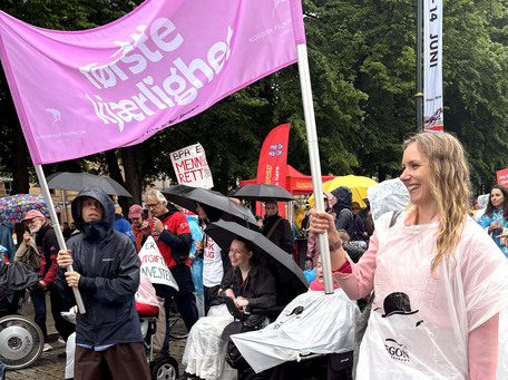 Funksjonshemmede og andre i regntøy i parade bak rosa banner med teksten 