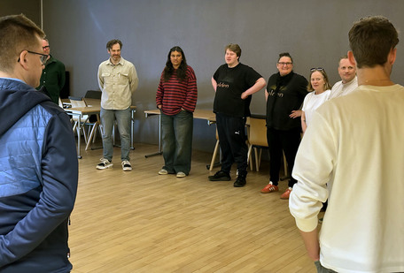 En stor gruppe mennesker står i ring i et seminarrom