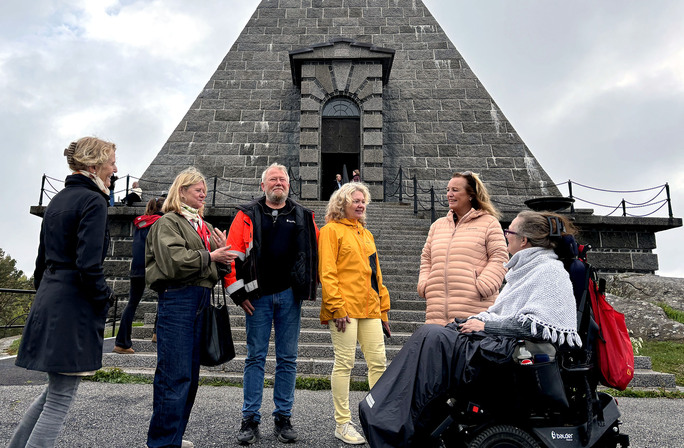 Seks mennesker, hvorav en kvinne i rullestol, foran den pyramideformede steinbygningen Minnehallen