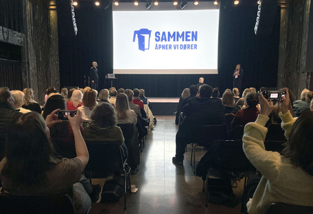 Full konfersansesal, storskjerm med bilde av innsamlingsbøsse og teksten 