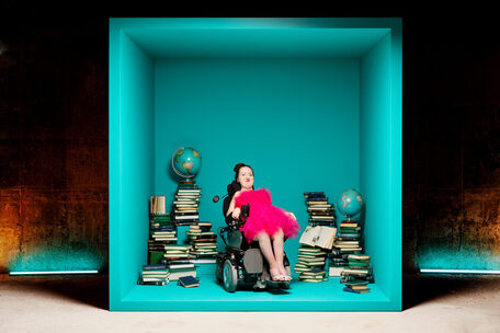 Ung kvinne med rosa kjole i rullestol inni en stor turkis boks med stabler av bøker toppet med globuser