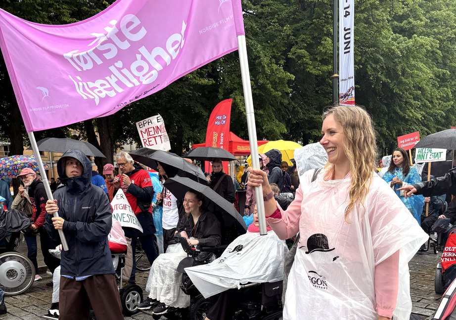 Funksjonshemmede og andre i regntøy i parade bak rosa banner med teksten 