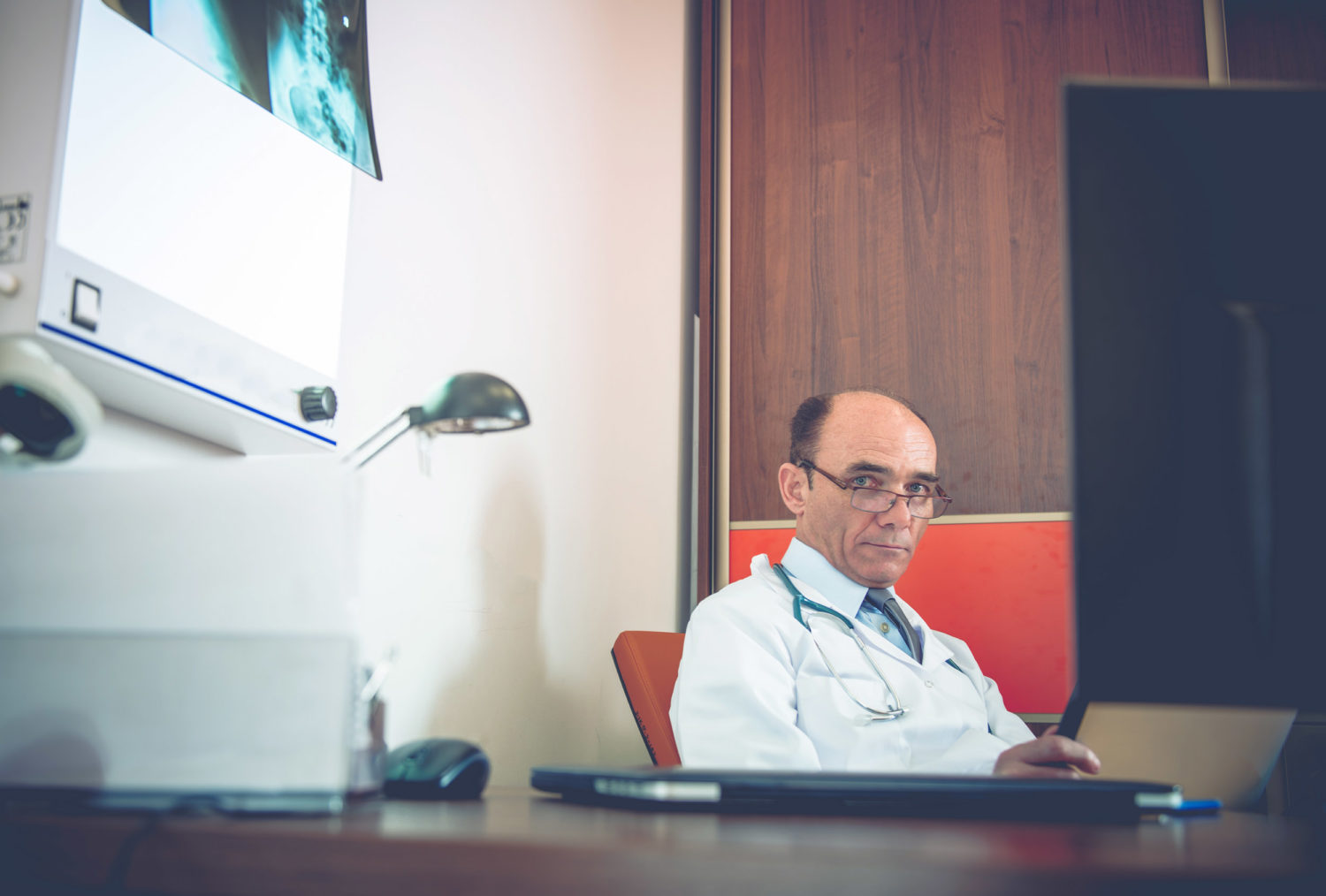 Foto av en alvorlig eldre lege i hvit frakk og stetoskop over skulderen, sittende bak et skrivebord.