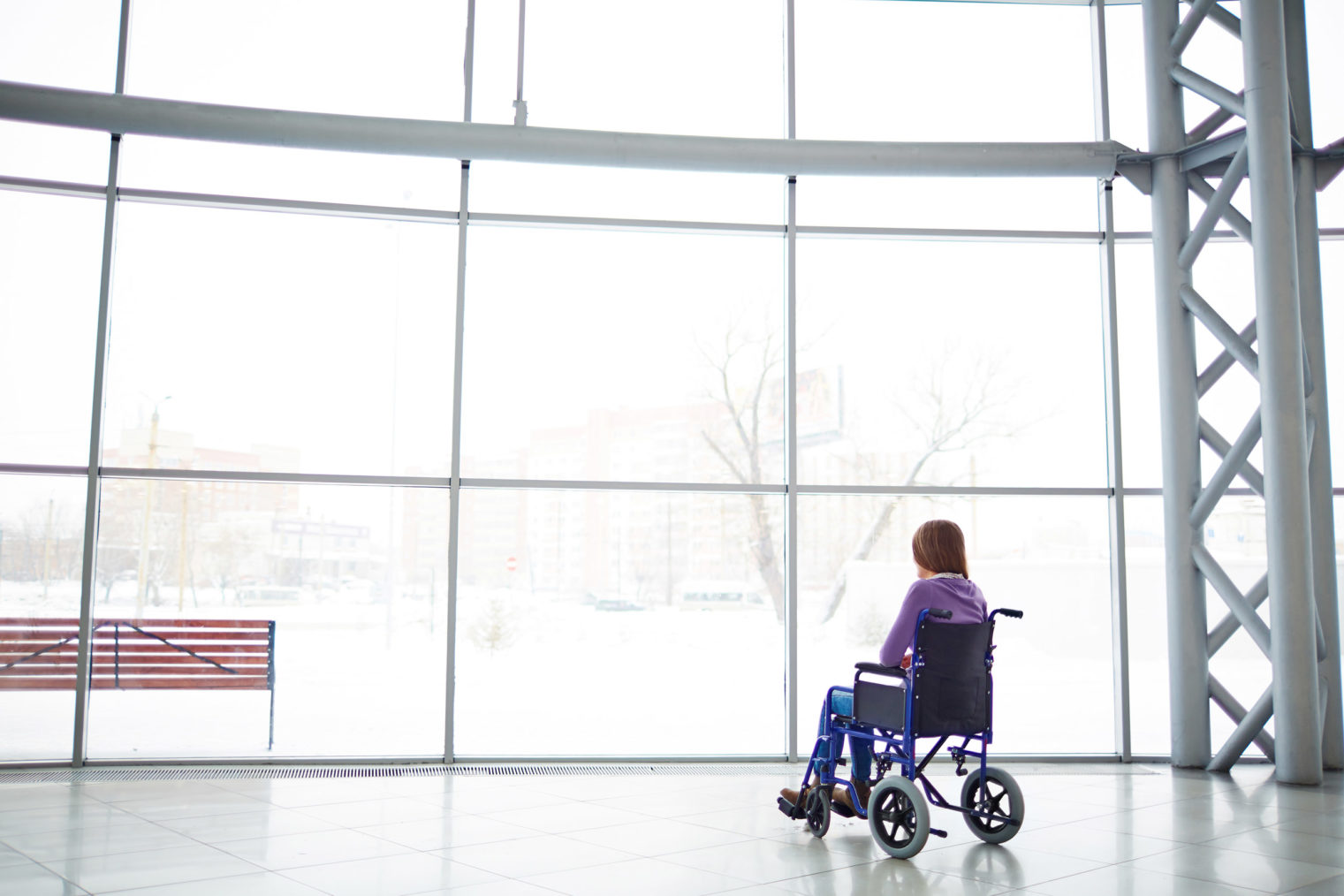 Bilde av kvinneskikkelse i rullestol på innsiden av en stor, lys vindusflate.