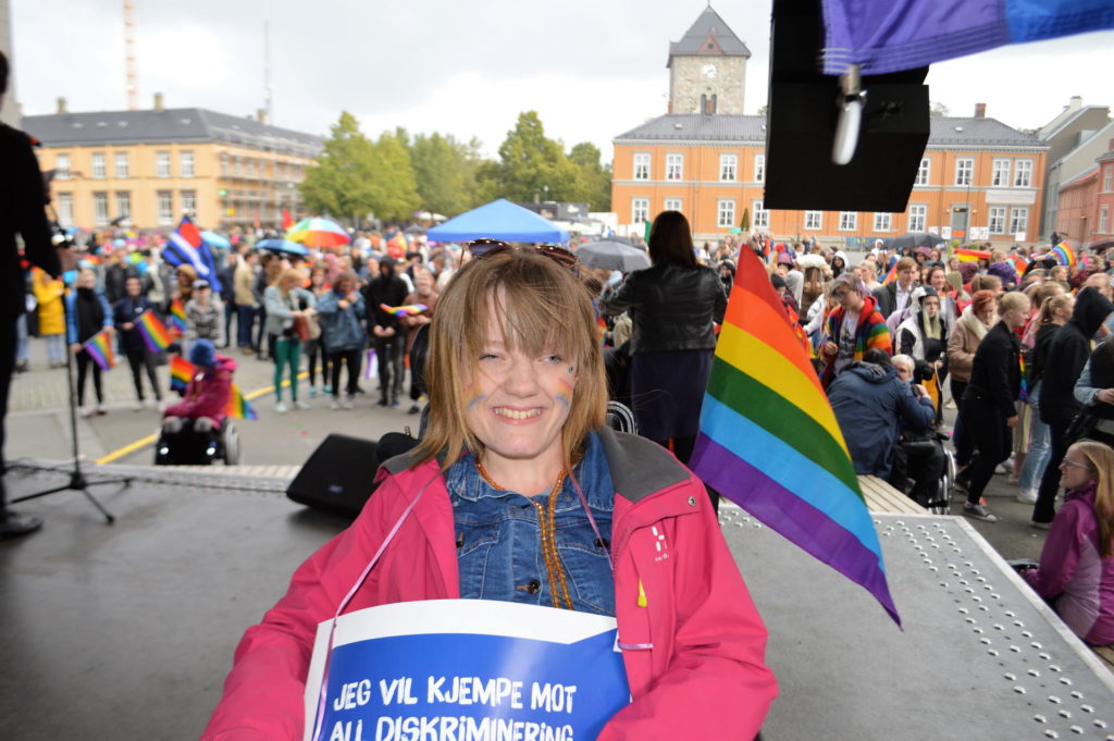 Marianne fotografert på scenen på Trondheim torg. Med regnbueflagg på rullestolen og malt i ansiktet.