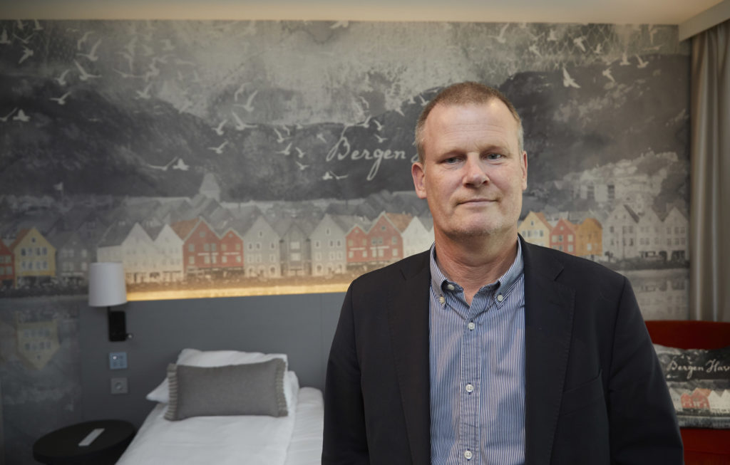 Berglund fotografert på et hotellrom med bilde av Bryggen i Bergen på veggen bak.