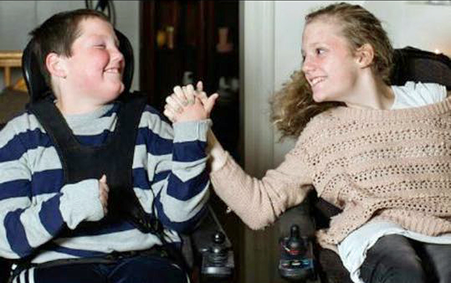 Bilde av Adrian og Marianne som sitter i hver sin rullestol, holder hender og smiler til hverandre.