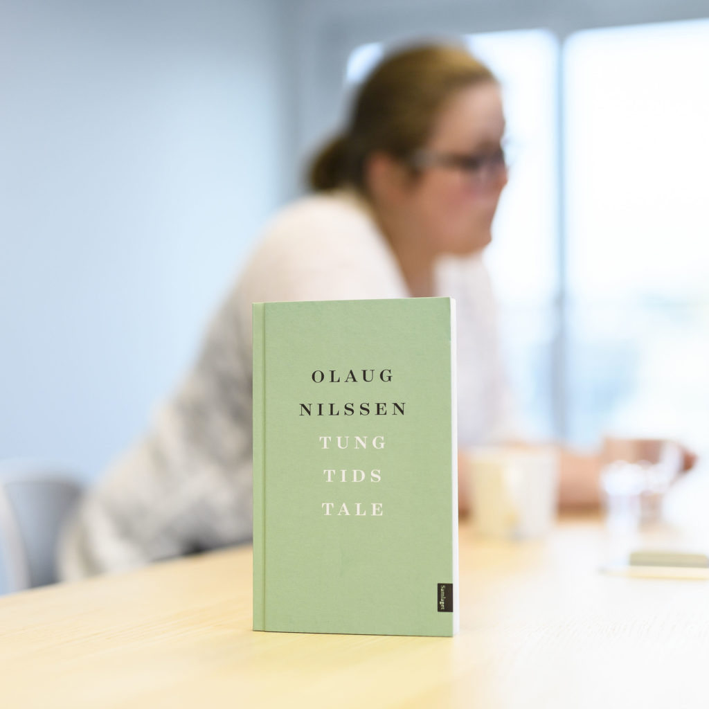 Olaug Nilssen sittende ute av fokus i bakgrunnen. Foran på bordet står boken hennes, «Tung tids tale».