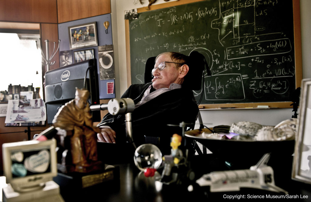 Hawking på kontoret med et rotete skrivebord i forgrunnen og en tavle på veggen bak.
