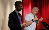 Eide og Astrup på et podium Eide står i lyset og holder en mikrofon, mens Astrup står i skyggen med foldede hender.