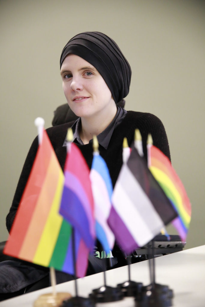 Ingrid Thunem fotografert ved et bord der det står små bordflagg som representerer ulike kjønnsidentiteter gjennom ulike varianter av regnbuetemaet.