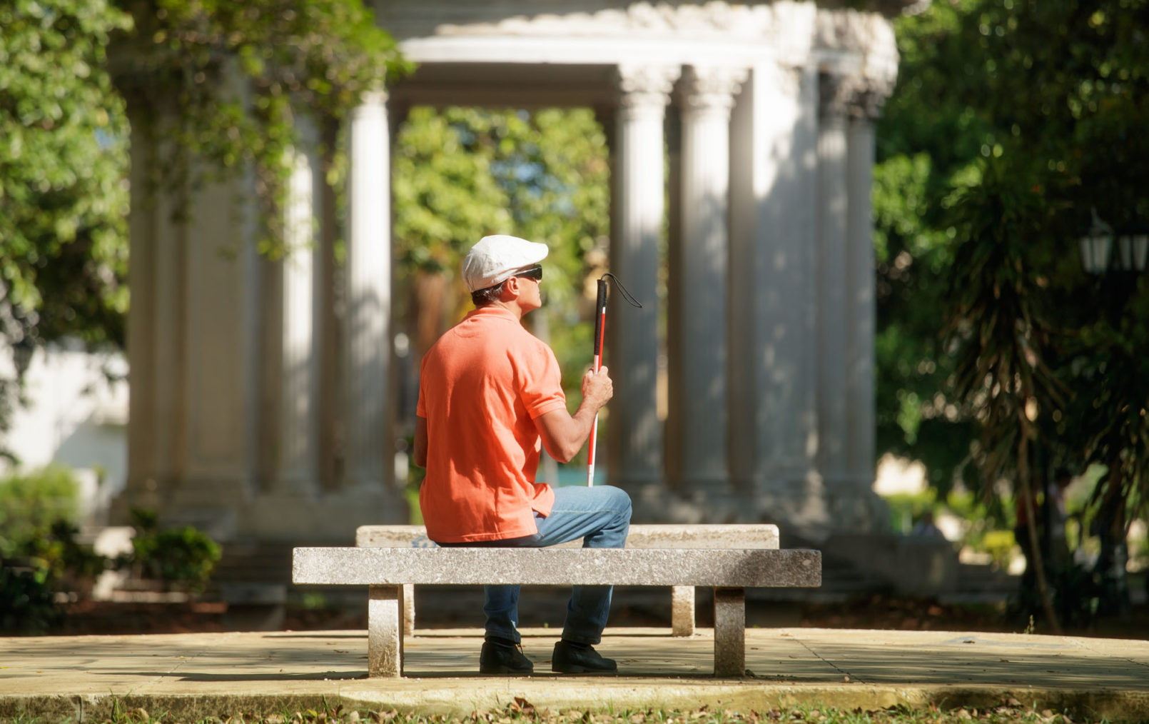 Bilde av mann med hvit stokk som sitter på en benk i en park.