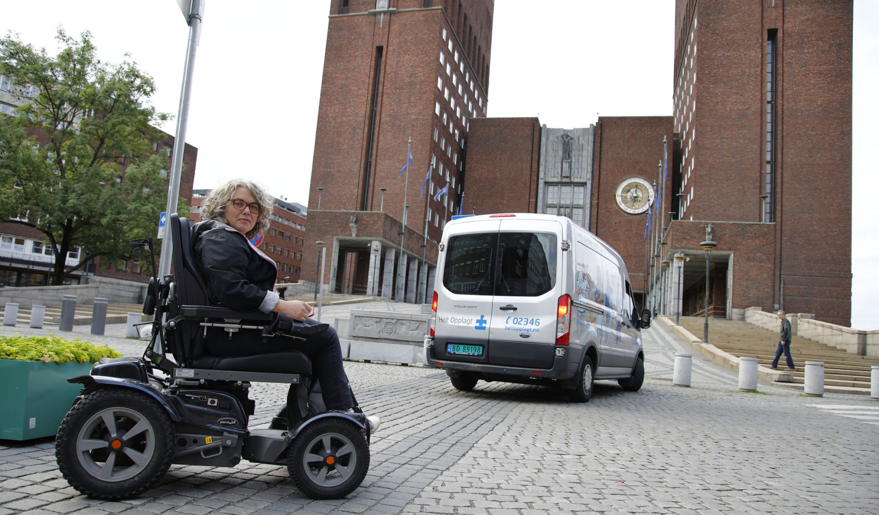 Magnhild Sørbotten i elektrisk rullestol utenfor rådhuset, mens varebil kjører inn mot inngangen.