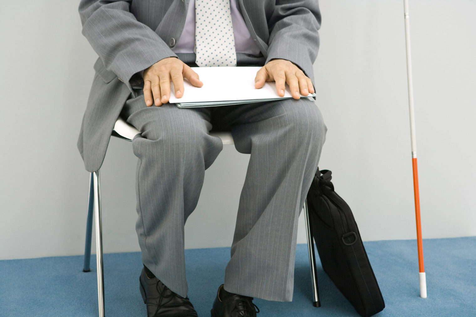 Nærbilde av mann i dress som sitter og venter med papirene sine i fanget. Ved siden av ham har han plassert en hvit stokk.