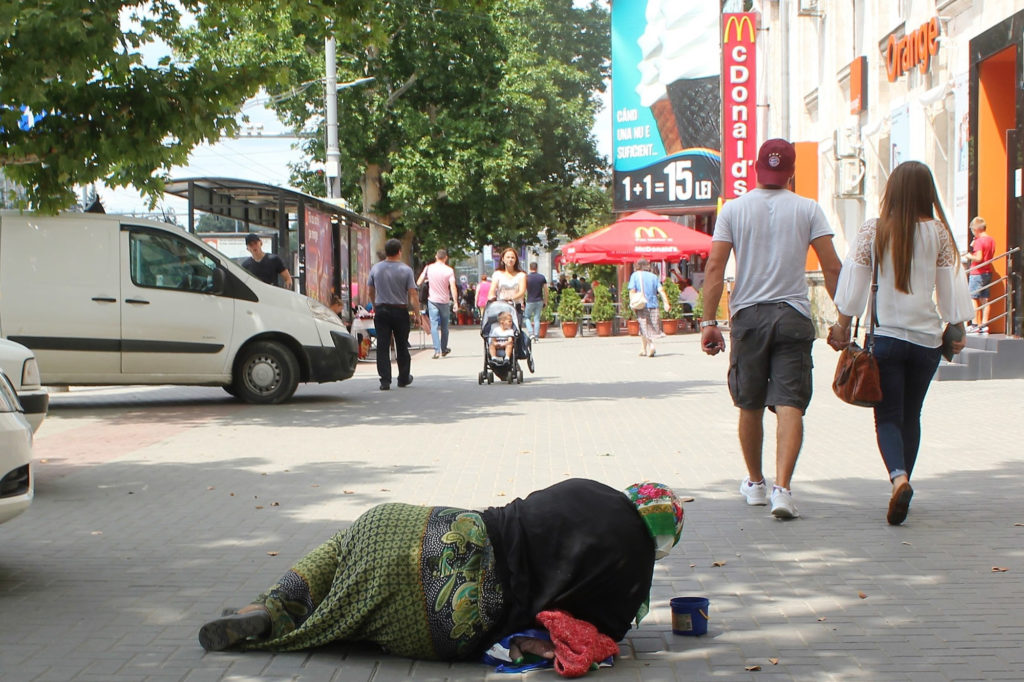 Eldre kvinne ligger på gaten og tigger, mens folk passerer forbi.
