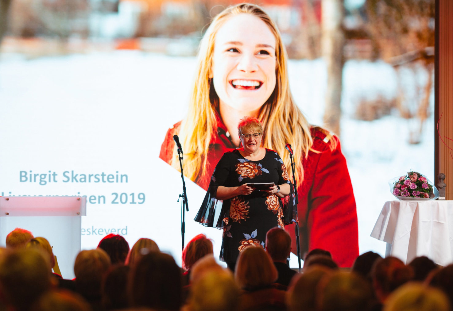 Grande snakker på scenen, med et stort bilde av Skarstein på skjerm i bakgrunnen.