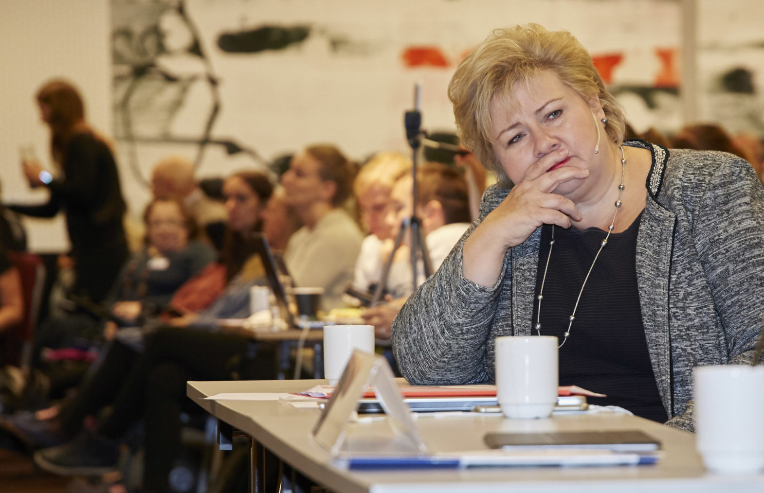 Erna lyttende i samtale ved et konferansebord. Masse mennesker i bakgrunnen.