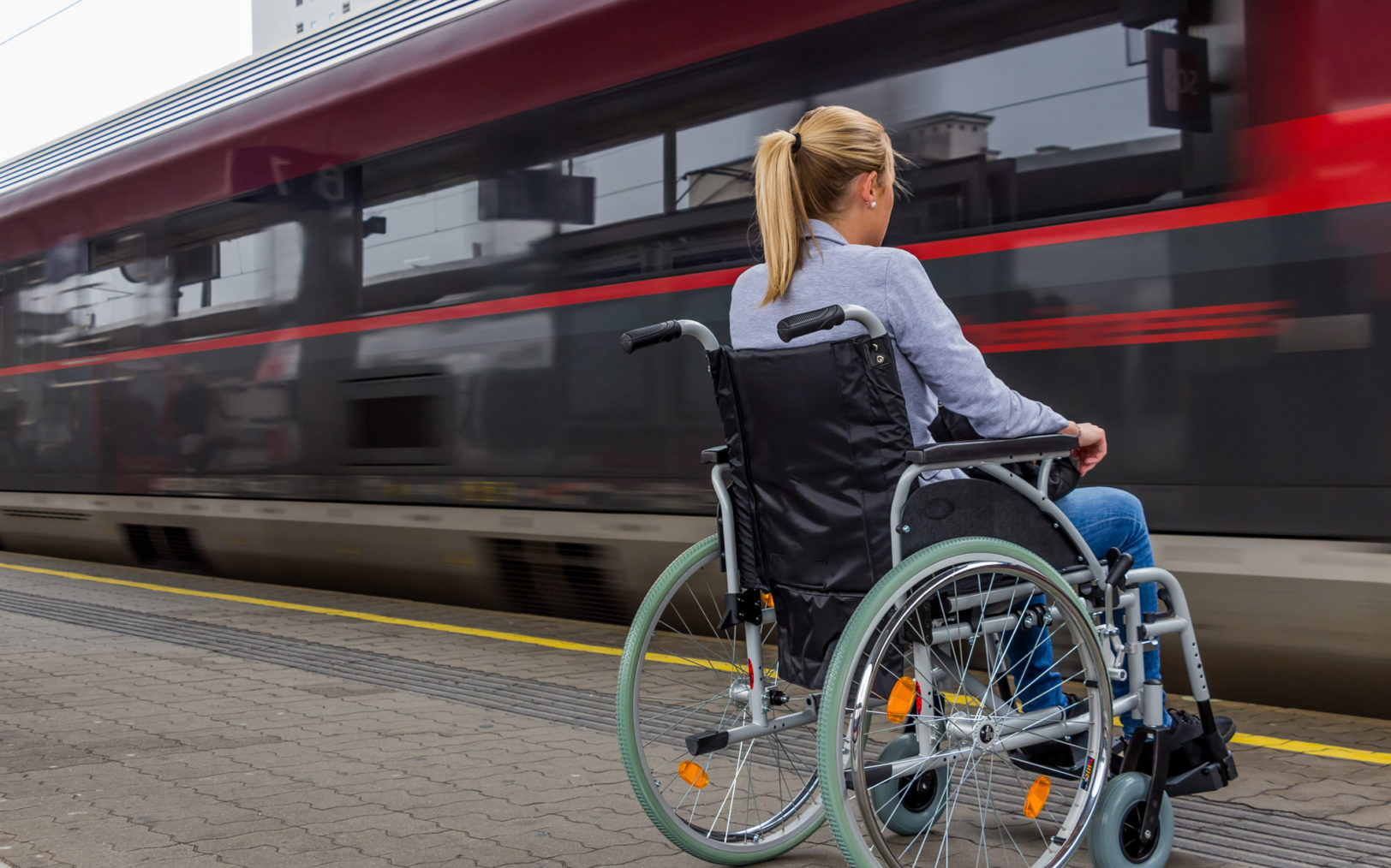 Kvinne i rullestol på jernbaneperrong mens tog passerer forbi.