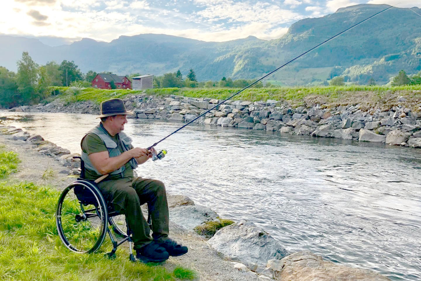Fjellheim sitter i rullestol ved en elv og fisker.