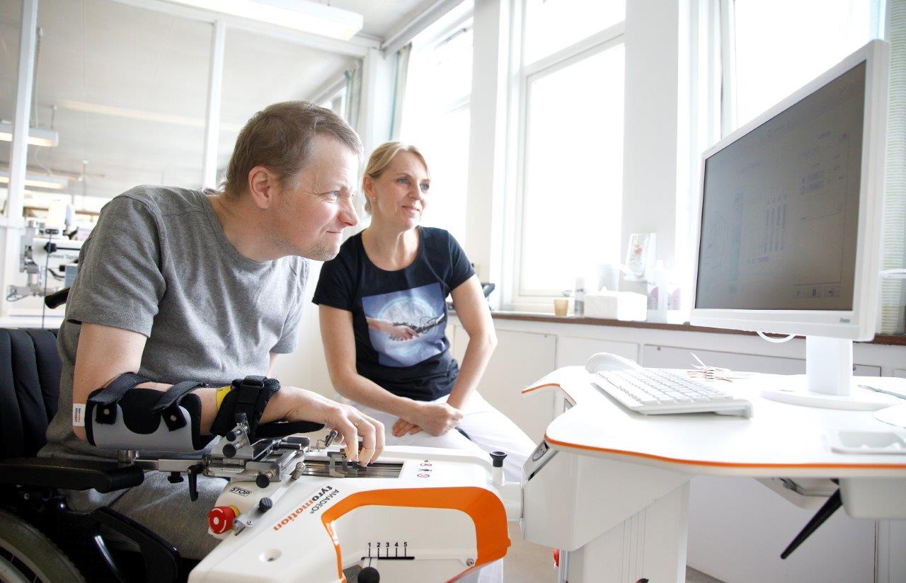 Stein Erik trener armen med maskinen, mens Linda ser på. Begge følger engasjert med på dataskjermen foran dem.