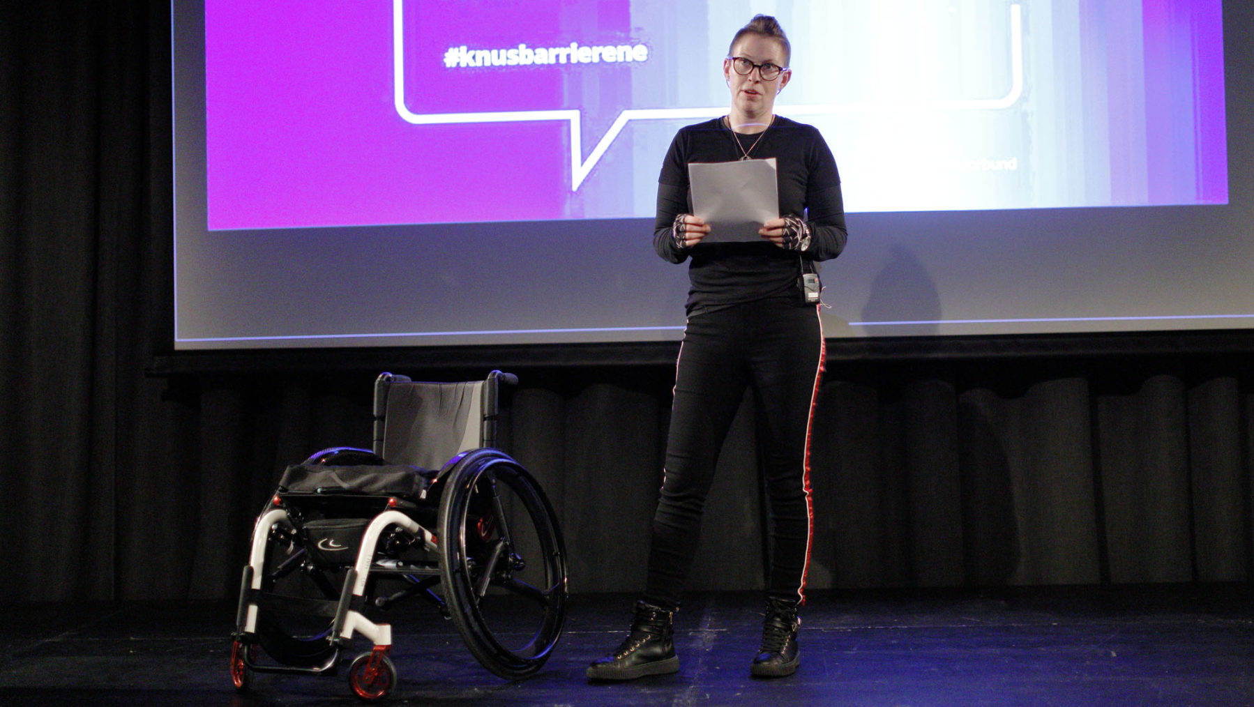 Ellen Merete Knutsen står på scenen med rullestolen ved siden av seg.
