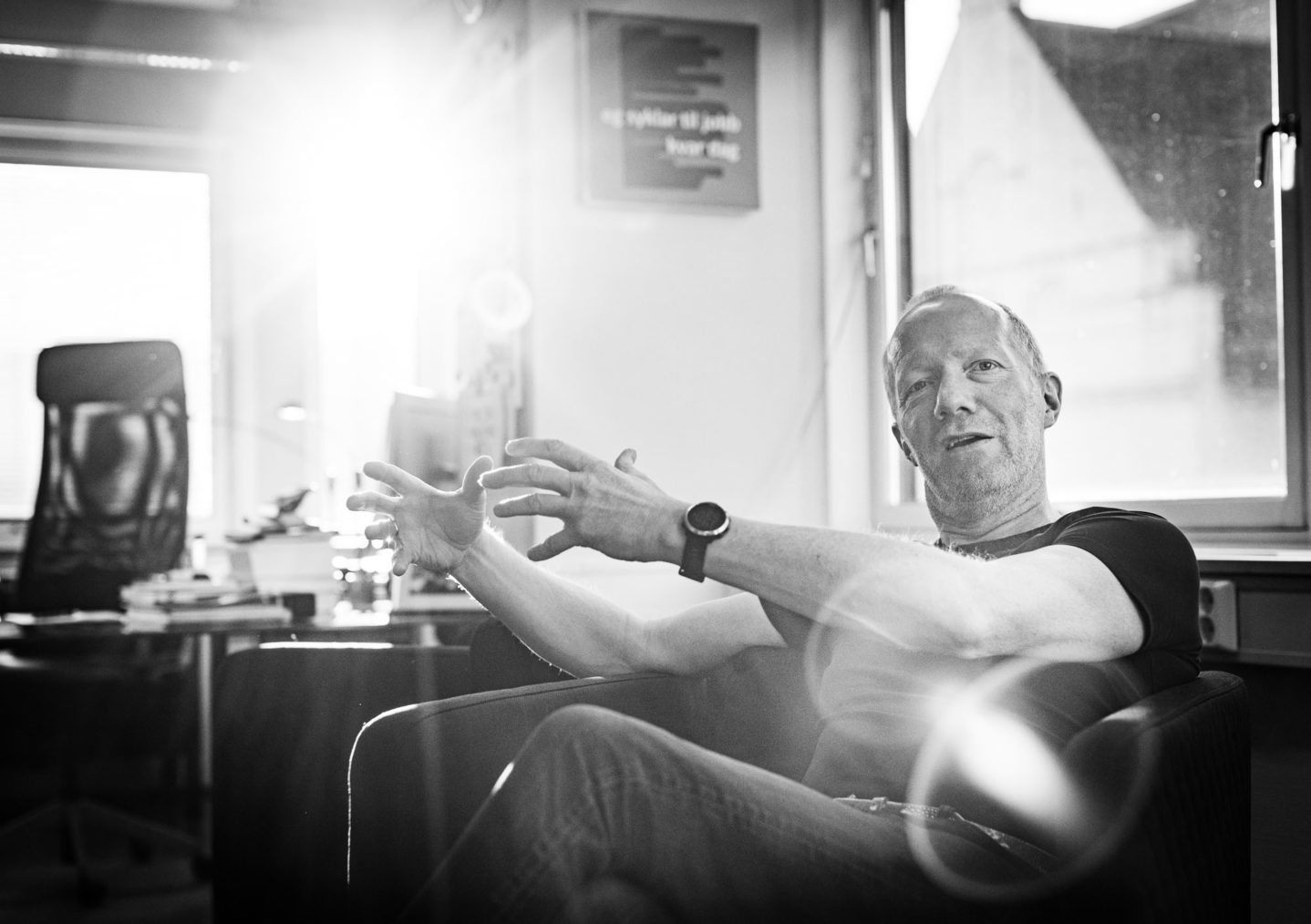 Arne Jensen fotografert mens han gestikulerer med hendene, som treffes av en solstråle som kommer inn gjennom vinduet bak ham.
