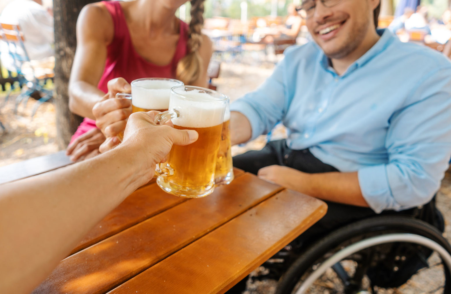 Tre venner rundt et bord skåler i øl. Den ene bruker rullestol.