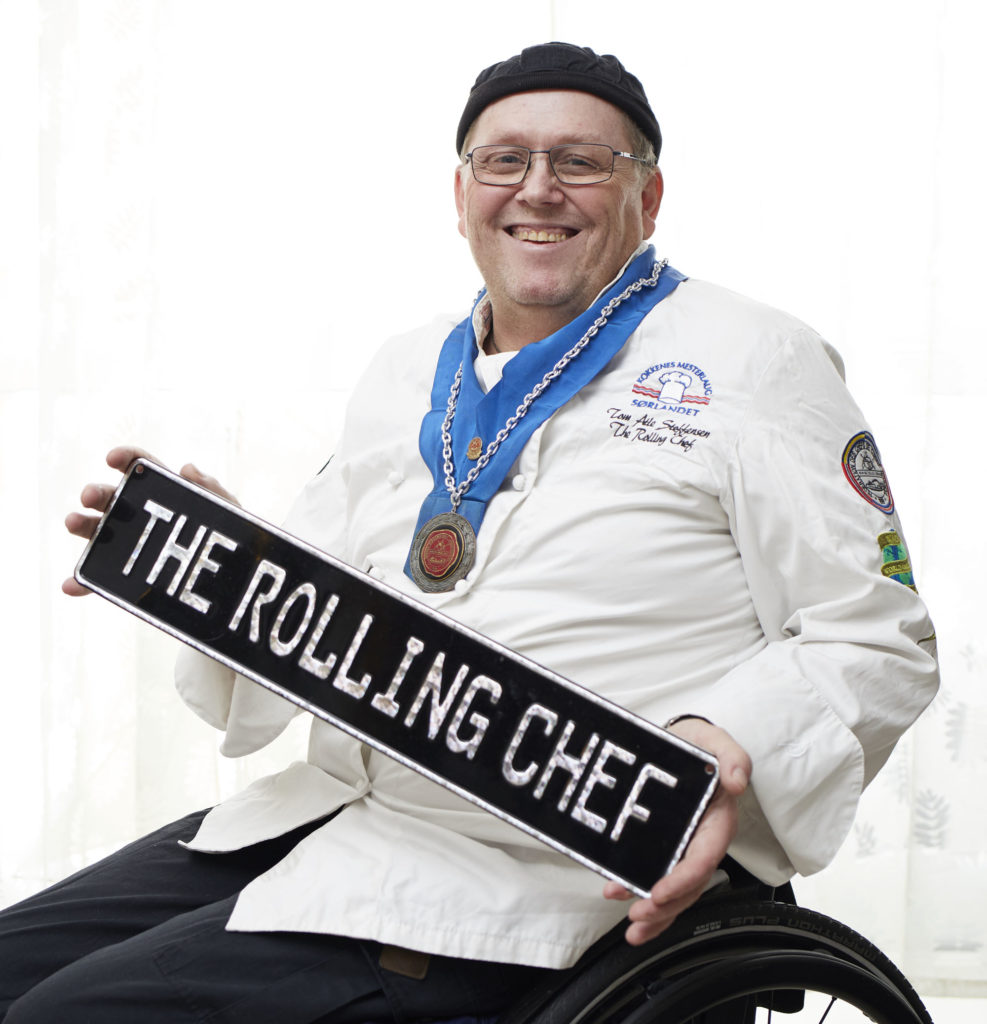Steffensen sitter i rullestol i kokkeklær og holder opp et skilt med «The Rolling Chef».