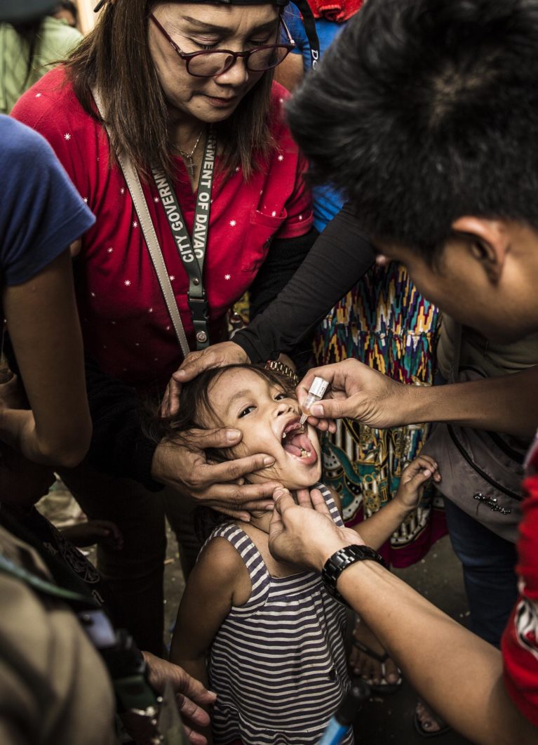 Helsearbeidere holder et barn som får vaksine dryppet i munnen.