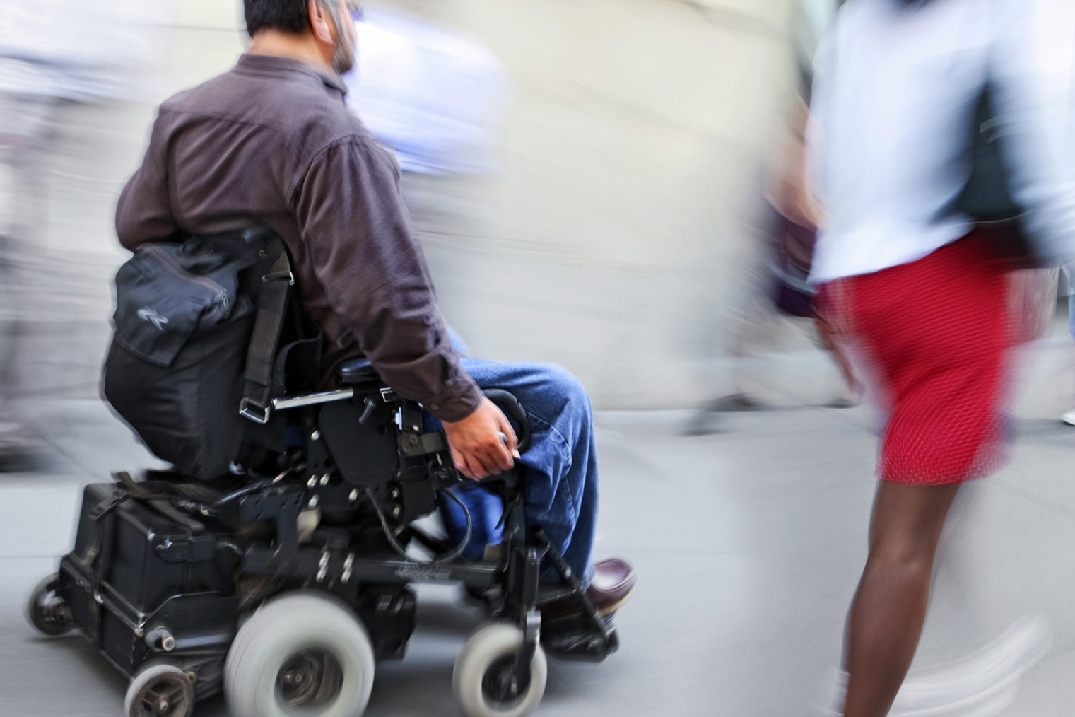Person i elektrisk rullestol passerer fotgjenger på fortau. Bildet har bevegelsesuskarphet for å gi illusjon av fart.