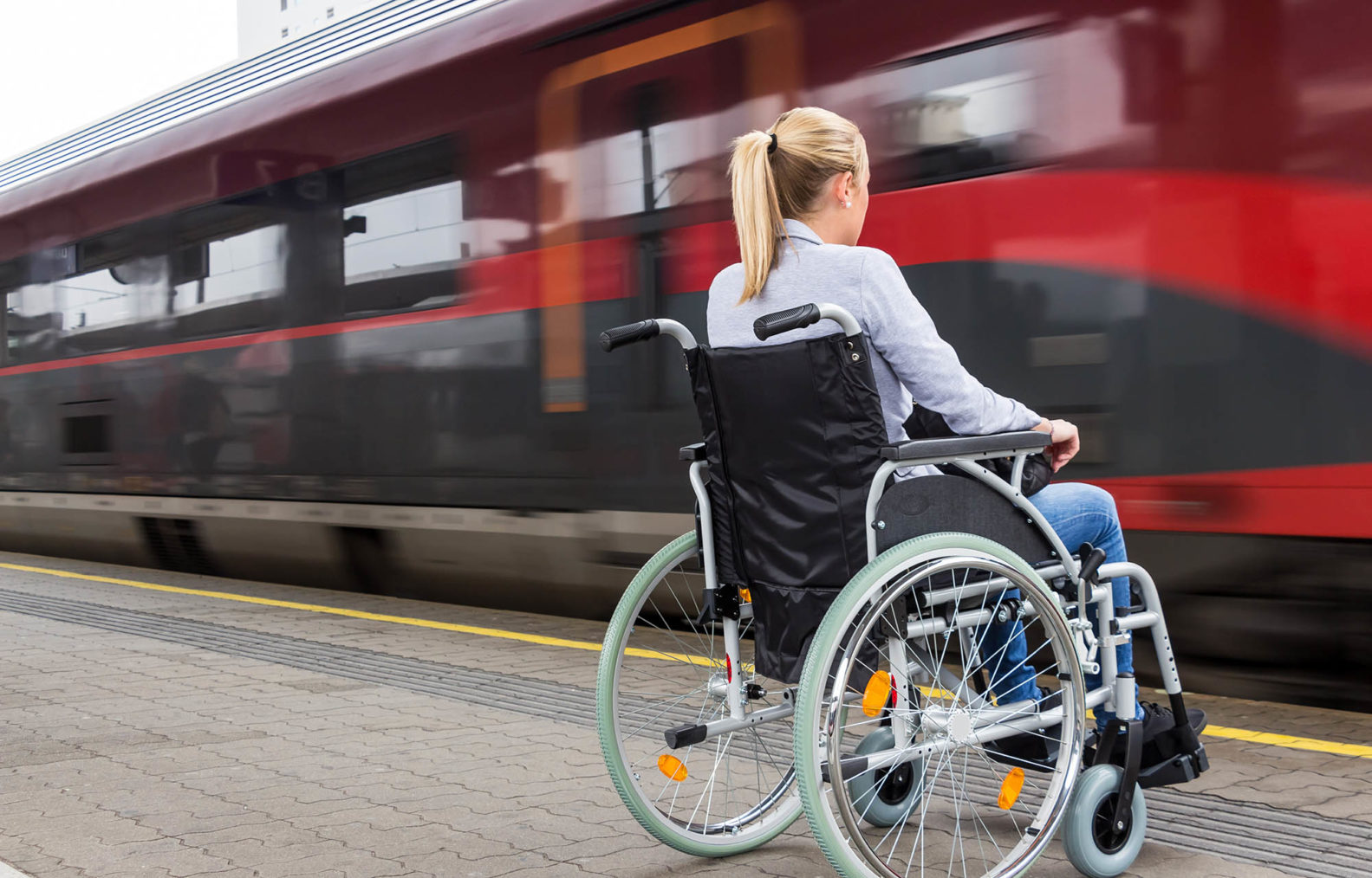 Ung kvinne i rullestol på perrong mens tog passerer forbi.