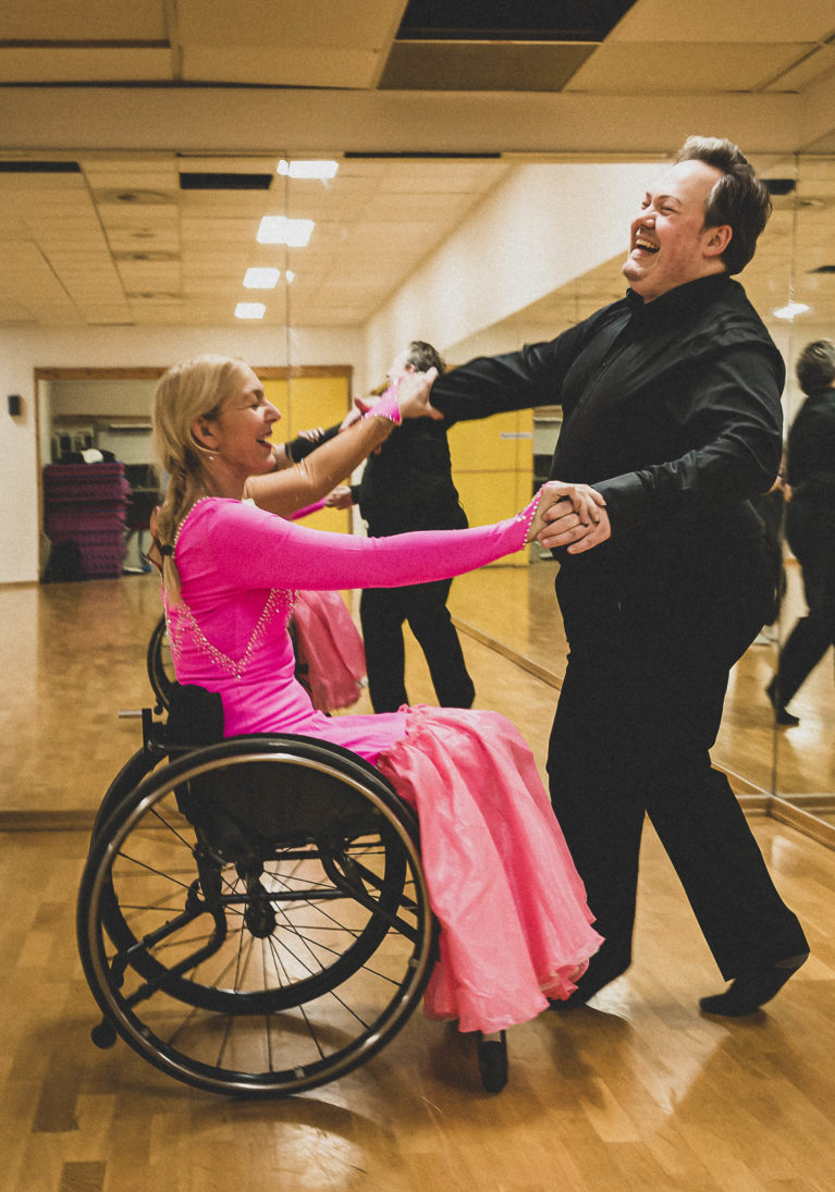 De to danser sammen. Hun i rullestol, han gående. Hun har på seg en rosa ballkjole med paljetter.