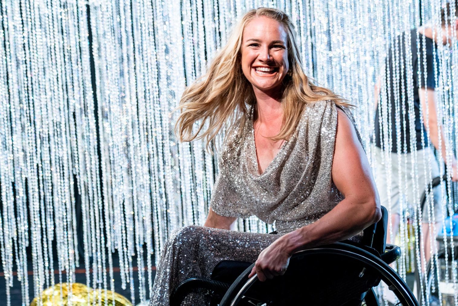 Birgit Skarstein i rullestol, med paljettkjole foran et glitterforheng på en scene.