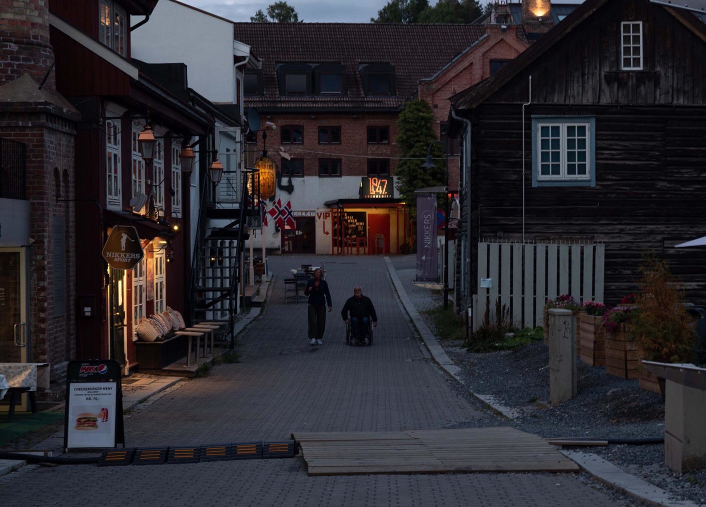 Kveldsbilde av gate med utesteder i Lillehammer. Midt i gaten er det en person i rullestol.