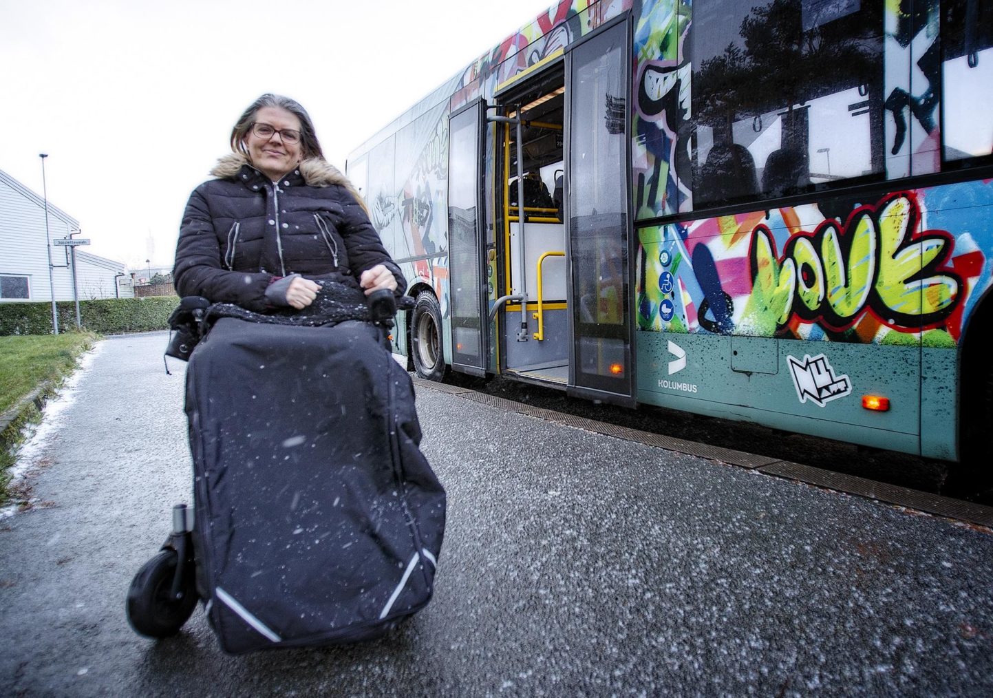 Gro Maren i rullestol på bussholdeplass ved siden av en buss.