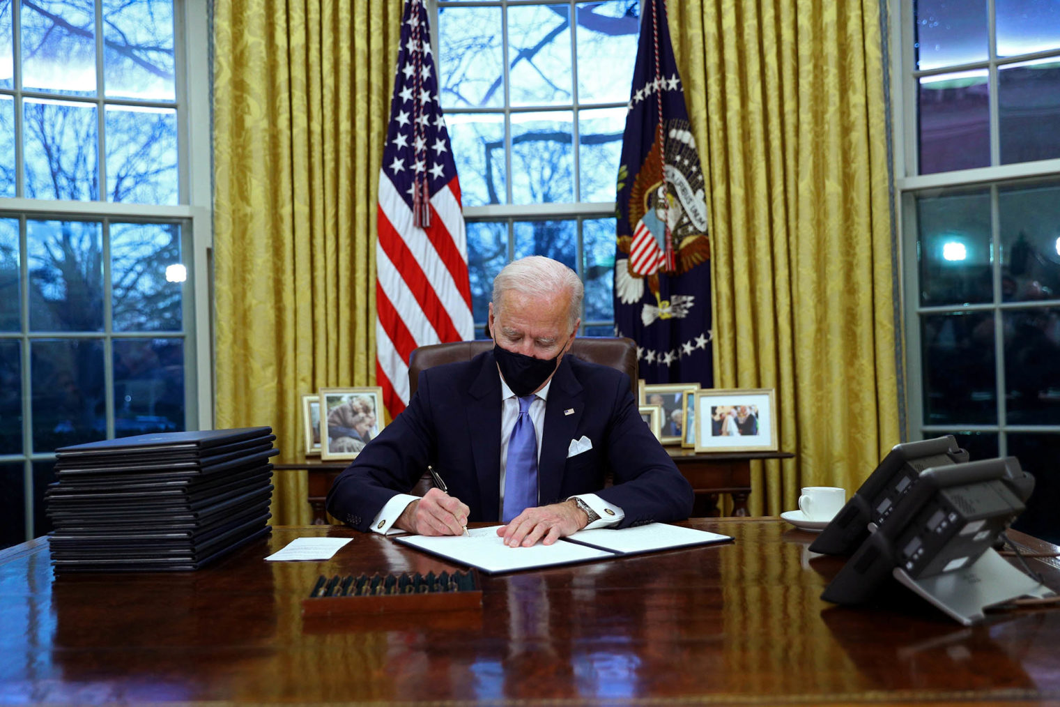 Joe Biden på presidentens kontor, med munnbind, sitter ved skrivebordet og signerer papirer.