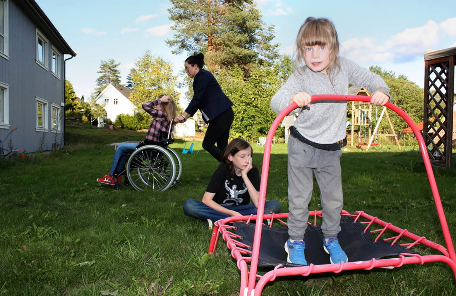 Familien sammen i hagen. Hege triller det ene barnet i rullestol, mens de to andre leker.