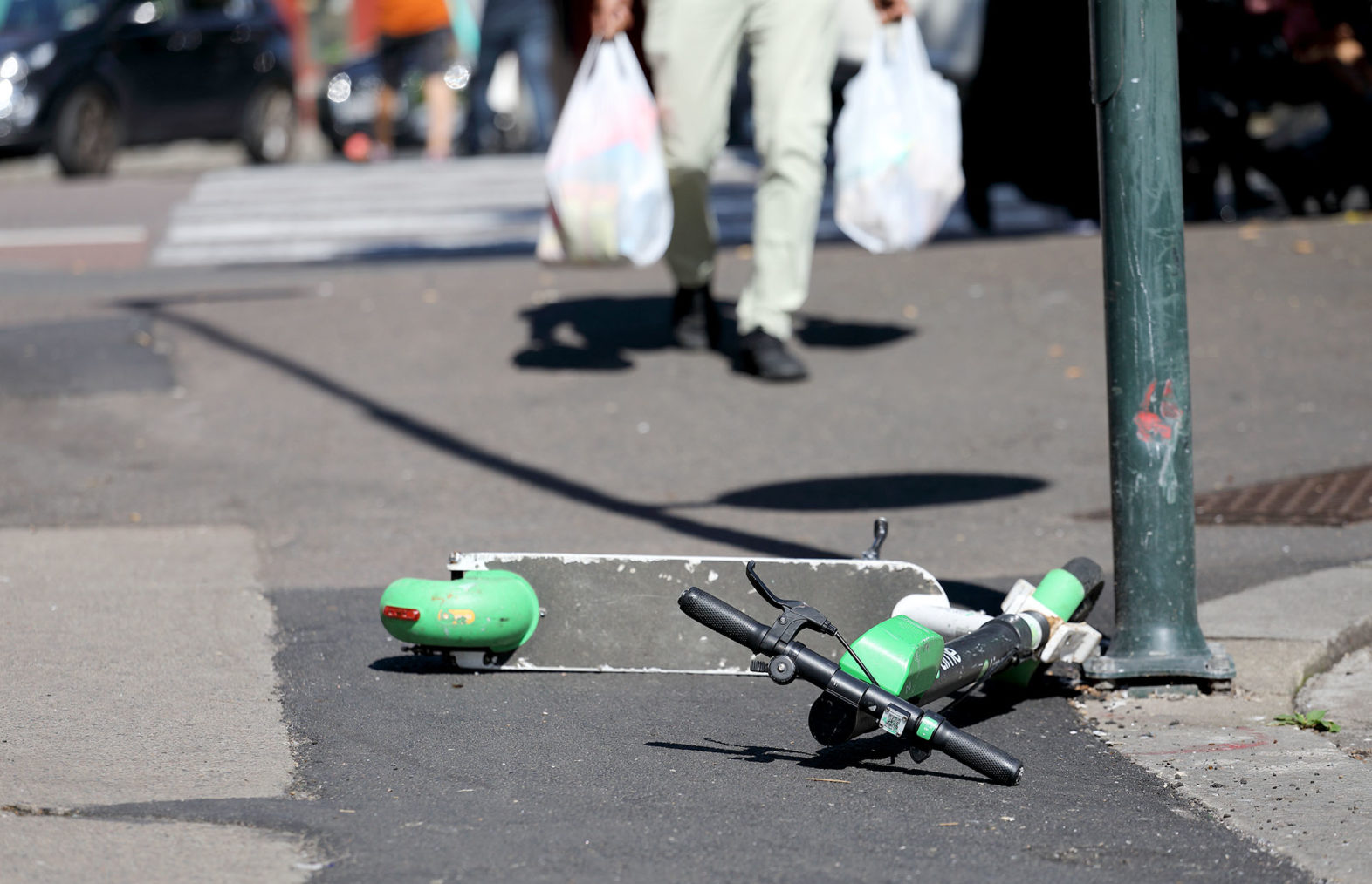 En el-sparkesykkel ligger på tvers av et fortau. I bakgrunnen kommer en fotgjenger med handleposer gående mot den.