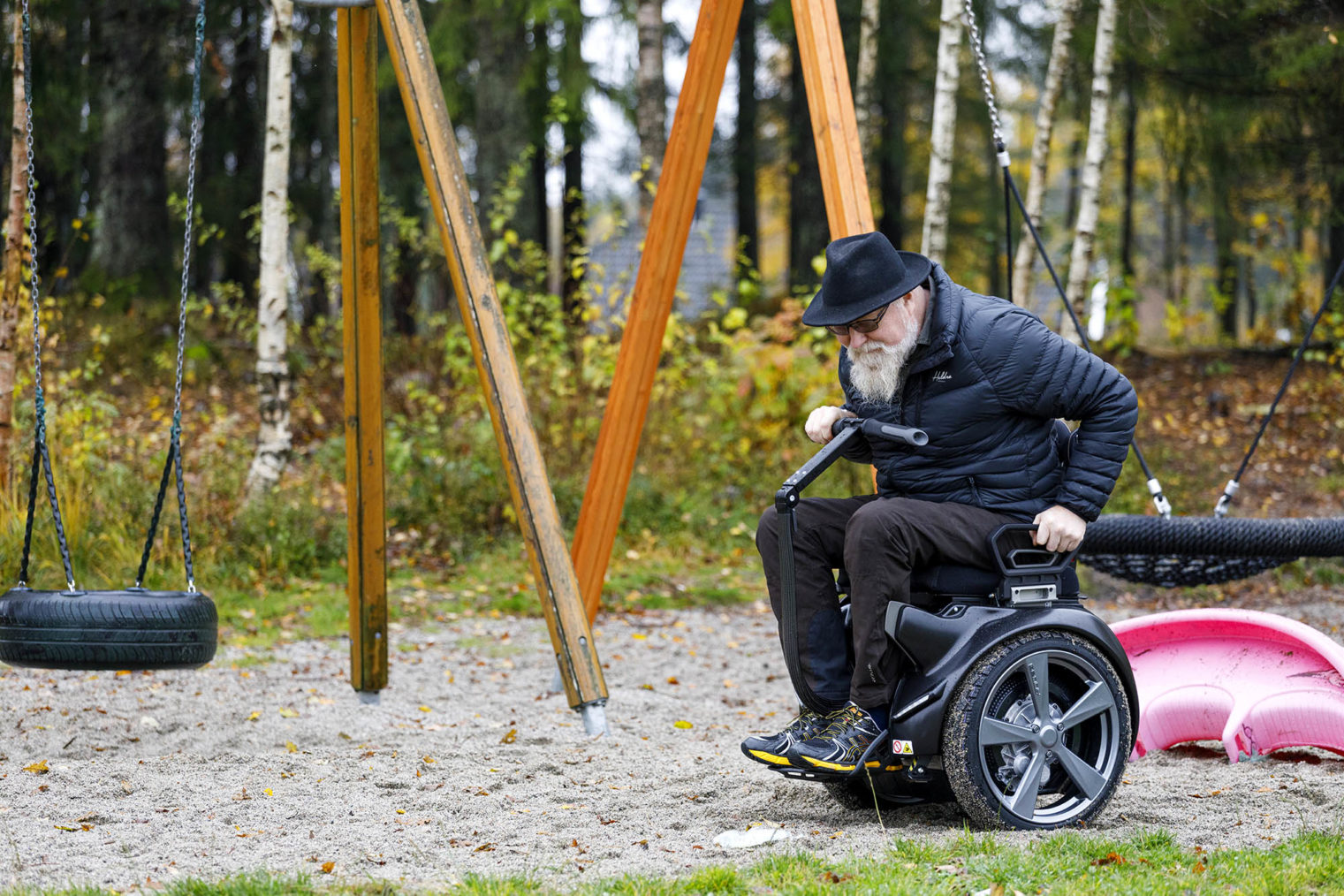 Langleite kjører rullestolen i sand, mellom huskestativer og lekeapparater.