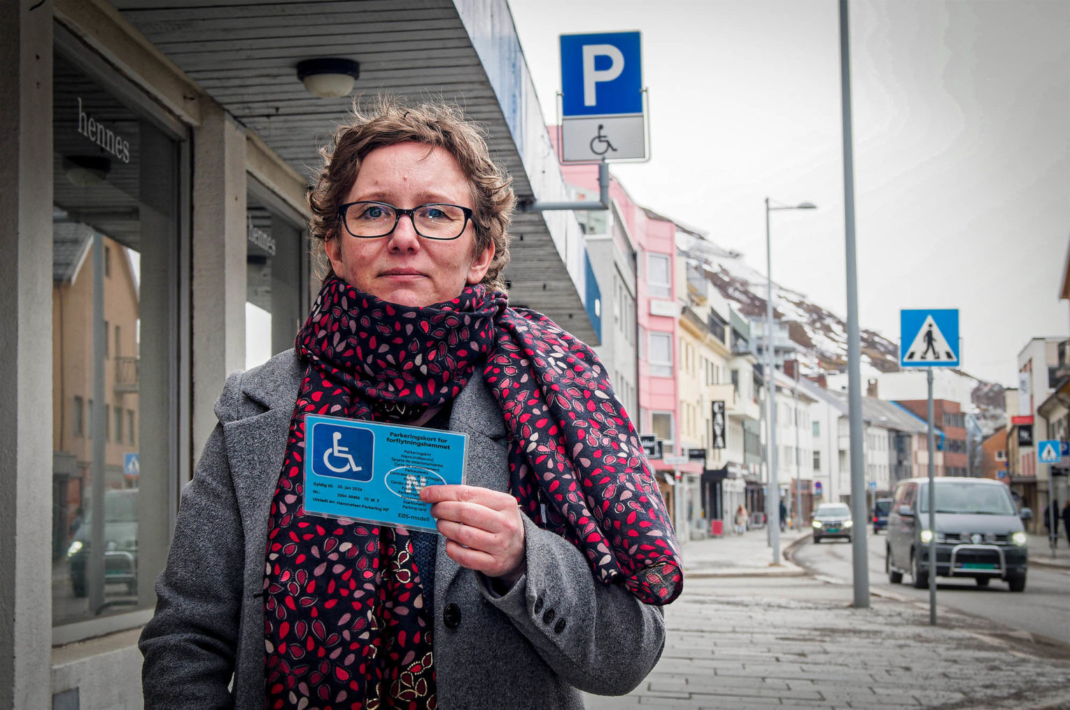 Lill Tove på gaten i Hammerfest holder opp parkeringsbeviset sitt. I bakgrunnen et parkeringsskilt med HC-symbol under.