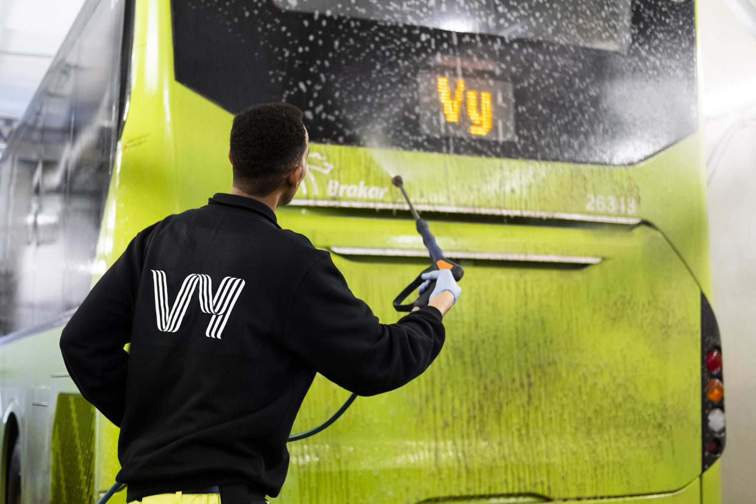Illustrasjonsbilde av en person med Vy-arbeidsklær som høytrykksspyler en Vy-buss.