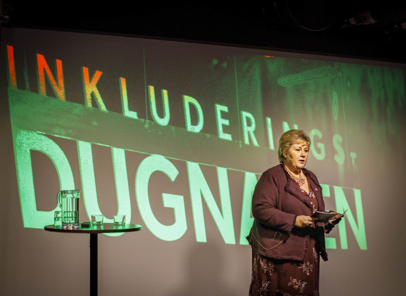 Erna Solberg holder et innlegg på et podium. «Inkluderingsdugnaden» er skrevet med store bokstaver på veggen bak henne.