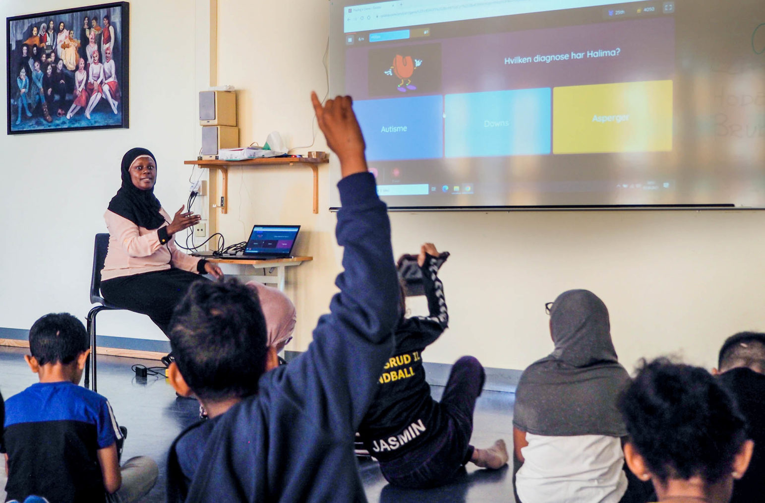 Bilde i klasserommet med elever på gulvet mens Faridah snakker. I forgrunnen rekker en gutt opp armen.
