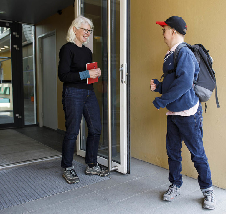 Hun står i døra og ønsker ham velkommen inn. Lars Jakob har ryggsekk på. De smiler til hverandre.