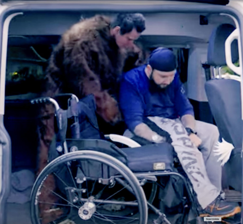 Mann i bjørnekostyme løfter og drar i Ole Rabbersvik for å få ham over i en rullestol.