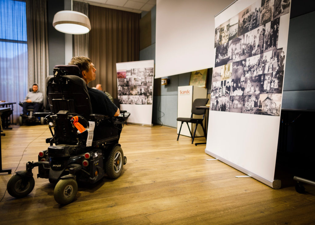 Kuløy i elektrisk rullestol betrakter gamle bilder fra Linde på veggen.