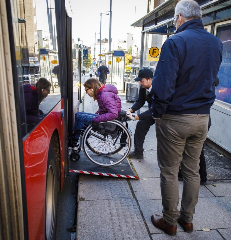 En sjåfør tar tak i Ingrids rullestol og dytter henne opp rampen inn på bussen.